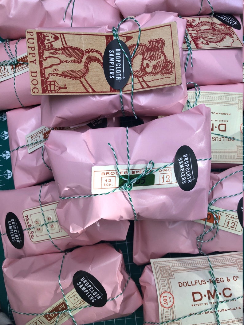 May include: A close-up of several pink packages wrapped with twine and labeled with "Dropcloth Samplers" and "D.M.C." The packages are arranged on a green grid surface.