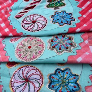 May include: A teal fabric with a white outline of a plate with embroidered Christmas-themed designs. The designs include a candy cane, a Christmas tree, a snowflake, and a peppermint swirl.