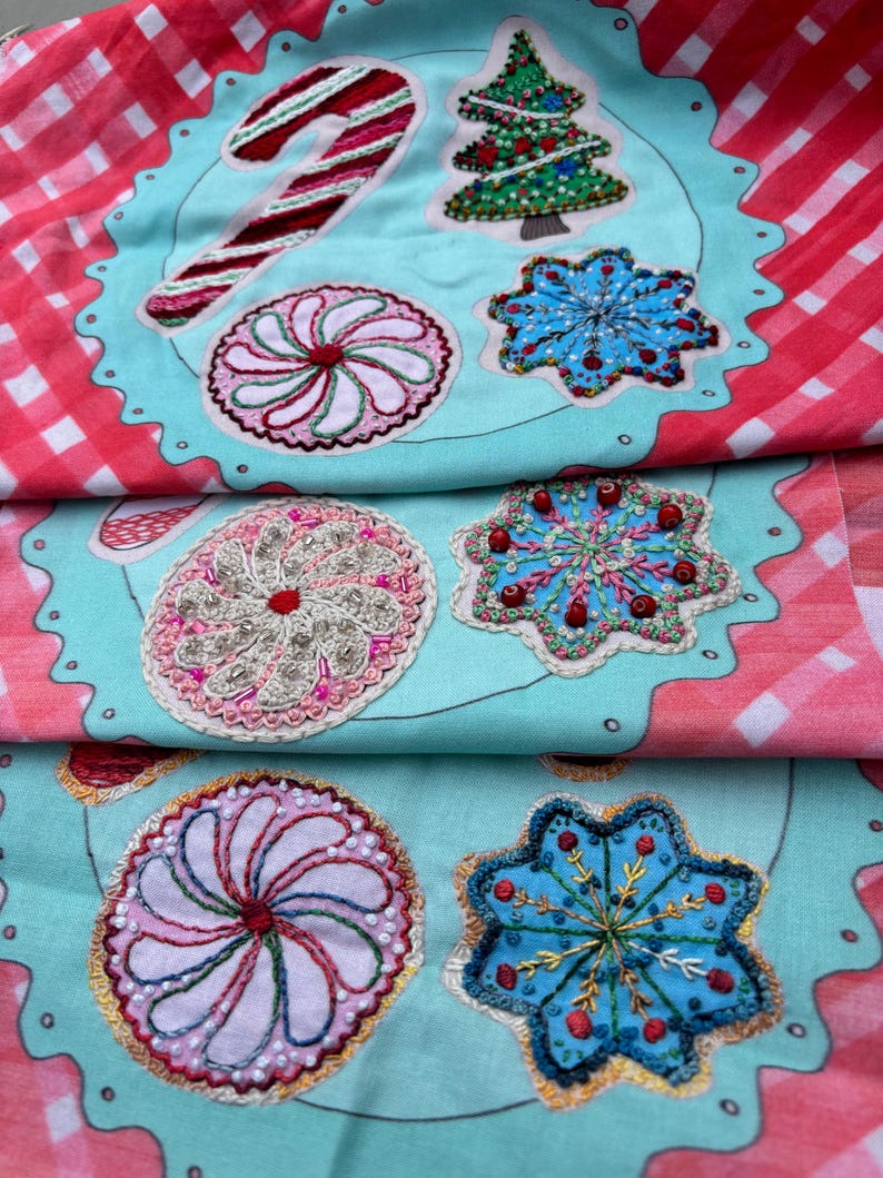 May include: A teal fabric with a white outline of a plate with embroidered Christmas-themed designs. The designs include a candy cane, a Christmas tree, a snowflake, and a peppermint swirl.
