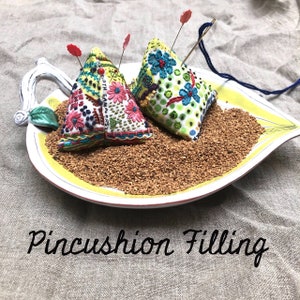 May include: Two colorful triangular pincushions filled with brown pincushion filling, sitting on a white and yellow leaf-shaped dish. The pincushions are decorated with embroidered flowers and dots. The text "Pincushion Filling" is written below the dish.