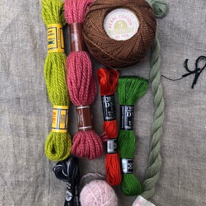 May include: Assortment of embroidery floss in various colors, including green, red, orange, and brown.  There are also two balls of yarn, one pink and one brown. The brown ball of yarn has the text "PEARL COTTON 5 JAP COATS 60 YDS" on the label.
