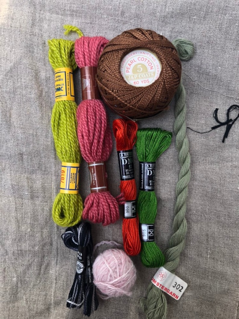 May include: Assortment of embroidery floss in various colors, including green, red, orange, and brown.  There are also two balls of yarn, one pink and one brown. The brown ball of yarn has the text "PEARL COTTON 5 JAP COATS 60 YDS" on the label.