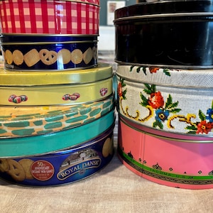 May include: A stack of vintage cookie tins in various colors and patterns. The tins include a red and white checkered pattern, a blue tin with cookies, a yellow tin with candy, and a pink tin with floral embroidery. The tins are stacked on a neutral surface.