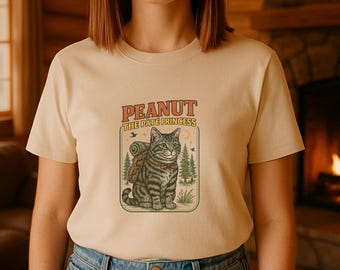 Könnte beinhalten: Beiges T-Shirt mit einer Cartoon-Katze mit Rucksack, umgeben von Bäumen und dem Text "PEANUT THE PATE PRINCESS". Die Katze ist grau und hat ein grünes Auge. Das Design befindet sich in einem rechteckigen Rahmen.