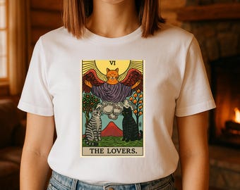May include: White t-shirt featuring a tarot card design with a cat theme. The card depicts a winged cat, two cats, trees, and the text "THE LOVERS." The design is in shades of yellow, orange, purple, and green.