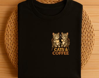 May include: Black t-shirt featuring an embroidered design of two cartoon cats seated and holding coffee cups. The text "CATS & COFFEE" is below the cats in a gold color. The t-shirt is folded on a woven, circular placemat.