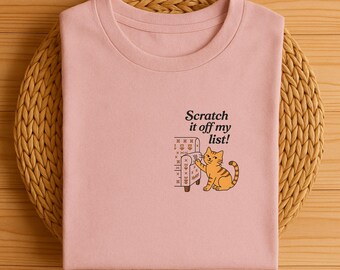 May include: A light pink t-shirt featuring a cartoon cat scratching a couch, with the text "Scratch it off my list!". The cat is orange with brown stripes. The couch is brown with a floral pattern.