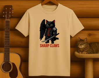 May include: A beige t-shirt featuring a black cat warrior with a sword, wings, and armor. The text "SHARP CLAWS" is printed in red below the cat illustration. The shirt is hanging on a wooden hanger.