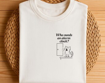 May include: A white t-shirt with a black graphic of a yawning cat next to a door. The text "Who needs an alarm clock?" is printed above the cat. The shirt is folded on a woven, circular placemat.