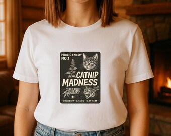 May include: White t-shirt with a graphic featuring a cat, catnip, and the text "CATNIP MADNESS". The design includes the words "PUBLIC ENEMY NO.1" and "WEED FROM THE DEVIL'S GARDEN!" in a vintage style.