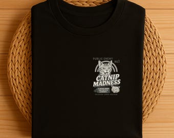 Könnte beinhalten: Schwarzes T-Shirt mit einer Grafik, die eine Katze und die Worte "CATNIP MADNESS" und "PUBLIC ENEMY NO.1" zeigt. Das Design beinhaltet eine Katzenillustration und zusätzlichen Text, alles in Weiß. Das T-Shirt liegt gefaltet auf einer gewebten, kreisförmigen Oberfläche.