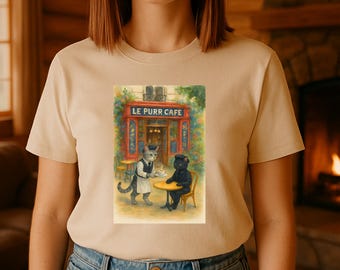 May include: Beige t-shirt featuring a whimsical illustration of a cat cafe scene. The artwork depicts a gray cat waiter serving coffee to a black cat at a table outside a cafe with the text "LE PURR CAFE" above the entrance.