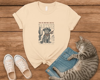May include: Cream-colored t-shirt with a graphic of a cat in a cowboy hat and bandana, the text "MEOWDY PARTNER" and "WHISKERS IN THE WIND". The shirt is paired with denim shorts and canvas sneakers. A tabby cat is sleeping nearby.