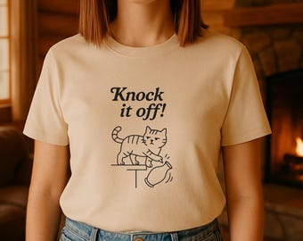 May include: Beige t-shirt with the phrase "Knock it off!" above a line drawing of a cat pushing a vase off a table. The cat is drawn in black, and the text is in a bold, black font.