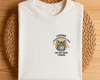 May include: A folded white t-shirt with a graphic of a gray cat inside a yellow circle. The text reads "I DESIRE UNCONDITIONAL LOVE ON MY OWN TERMS". The shirt is on a woven, circular placemat.