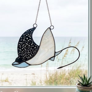 Puede incluir: Un atrapasoles de vidrieras con forma de mantarraya, que presenta un ala negra con motas blancas y un ala blanca de textura lisa. La pieza está suspendida por una cadena de metal e incluye una cola de metal larga, fina y curvada.