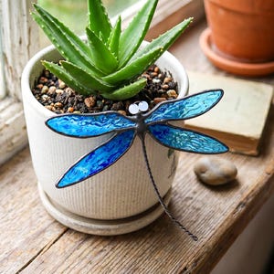 Può includere: Un ornamento a forma di libellula in vetro colorato con ali blu testurizzate e occhi a perla bianchi, fissato al lato di un vaso in ceramica bianca contenente una pianta di aloe verde. La libellula presenta saldature in metallo scuro.