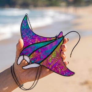 May include: A stained glass manta ray ornament featuring a vibrant galaxy-inspired pattern with swirls of purple, blue, and green, accented by white speckles. The piece is constructed with dark metal solder lines and includes a thin black hanging chain.