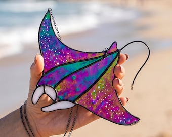 May include: A stained glass manta ray ornament featuring a vibrant galaxy-inspired pattern with swirls of purple, blue, and green, accented by white speckles. The piece is constructed with dark metal solder lines and includes a thin black hanging chain.