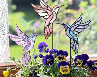 Può includere: Tre picchetti da giardino a forma di colibrì in vetro colorato in un vaso di terracotta con viole del pensiero viola e gialle. I colibrì sono rosa, viola e blu con picchetti in metallo nero. Il vaso è su un davanzale di legno.