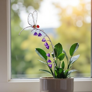 May include: A decorative metal dragonfly and purple flower plant stake in a beige ceramic pot. The dragonfly has clear wings and a red bead head. The plant is on a windowsill with a notebook and glasses.