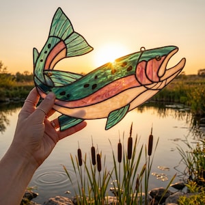 A stained glass fish suncatcher featuring translucent green, pink, and white glass panels held together by dark metal solder lines. The fish has a curved body shape, a detailed tail fin, and small black spots along its upper back.