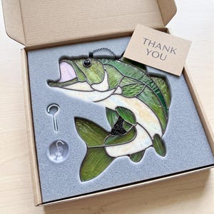 May include: A stained glass fish suncatcher in shades of green and white with a black eye, packaged in a cardboard box with gray foam inserts. Included are a metal screw hook, a clear suction cup, and a tan card with the text THANK YOU.