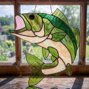 May include: A stained glass suncatcher shaped like a leaping fish, featuring textured green and cream-colored glass panels joined by dark metal solder lines. The fish has an open mouth with a pale pink interior and a single dark glass eye.