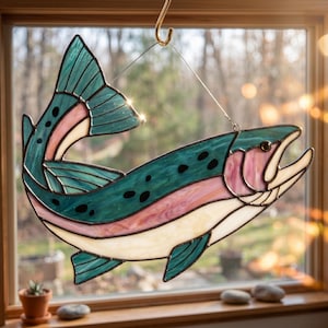 May include: A stained glass suncatcher shaped like a trout, featuring teal, pink, and cream-colored glass panels joined by dark metal solder lines. The fish is depicted in a curved, leaping pose and is suspended by a thin wire from a metal hook.