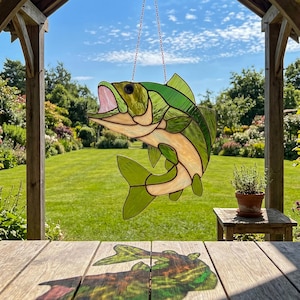 May include: A stained glass suncatcher shaped like a leaping fish with green and cream-colored glass panels, a pink open mouth, and a black eye. The fish is suspended by a thin metal chain, casting a colorful shadow on a wooden surface below.