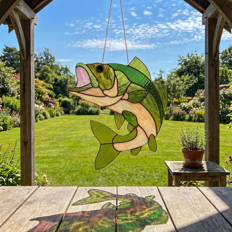 May include: A stained glass suncatcher shaped like a leaping fish with green and cream-colored glass panels, a pink open mouth, and a black eye. The fish is suspended by a thin metal chain, casting a colorful shadow on a wooden surface below.