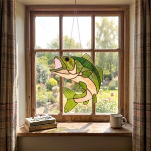 May include: A stained glass suncatcher shaped like a leaping fish with green and cream-colored glass panels, dark solder lines, and a pink open mouth, suspended by a thin metal chain in front of a window pane with a view of a garden.