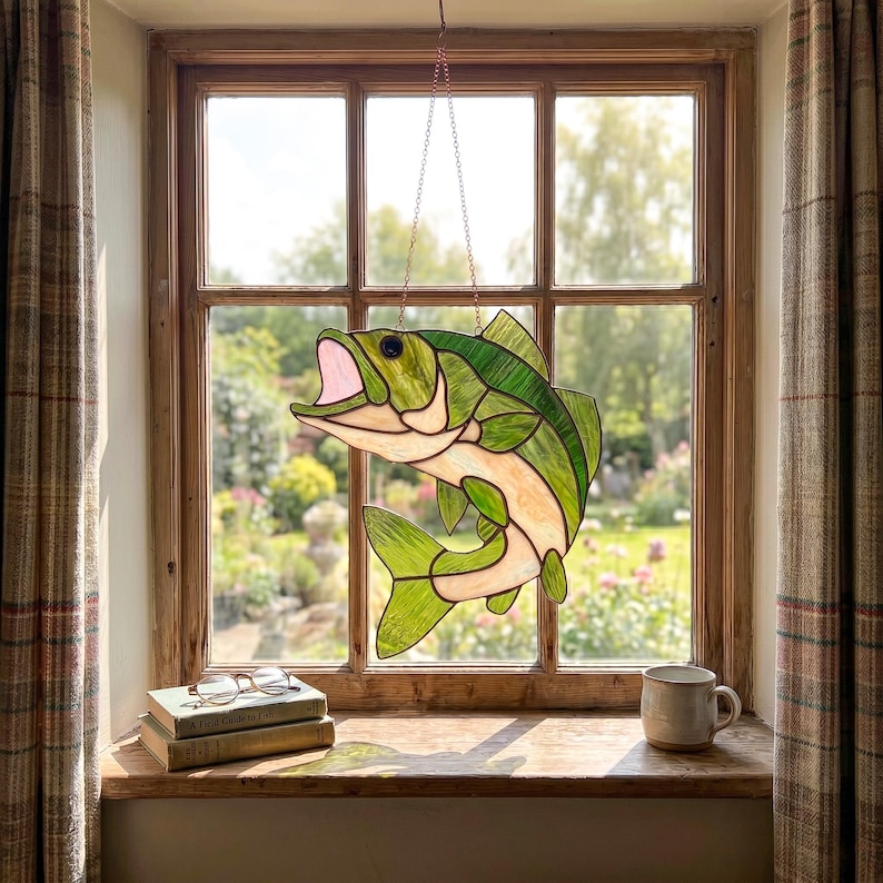 May include: A stained glass suncatcher shaped like a leaping fish with green and cream-colored glass panels, dark solder lines, and a pink open mouth, suspended by a thin metal chain in front of a window pane with a view of a garden.
