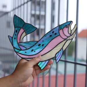 May include: A stained glass fish suncatcher featuring iridescent teal, pink, and cream-colored glass panels joined by dark metal solder lines. The fish is depicted in a curved, leaping posture with a detailed tail fin and dark spots.