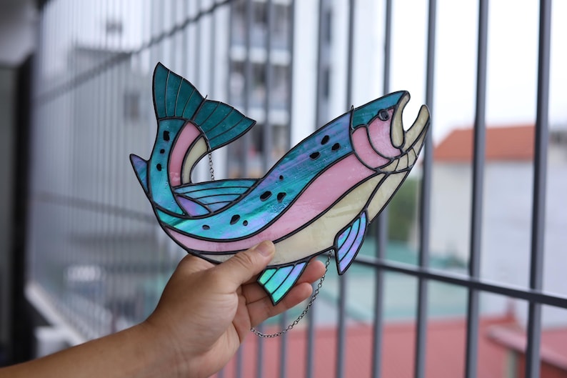 May include: A stained glass fish suncatcher featuring iridescent teal, pink, and cream-colored glass panels joined by dark metal solder lines. The fish is depicted in a curved, leaping posture with a detailed tail fin and dark spots.