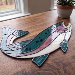 May include: A stained glass fish suncatcher featuring a curved body with deep teal, iridescent pink, and translucent white glass segments. The piece is constructed with dark metal solder lines and includes small black glass spots along the back.