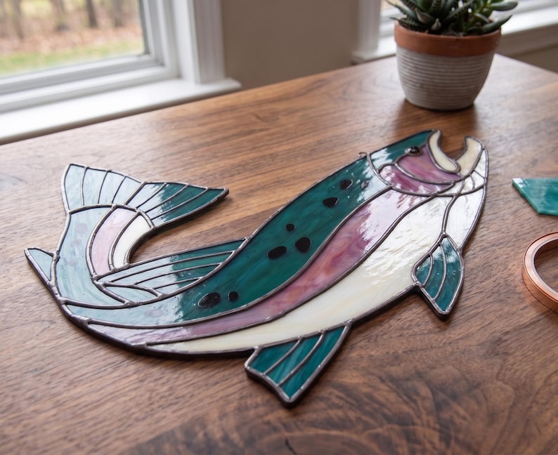 May include: A stained glass fish suncatcher featuring a curved body with deep teal, iridescent pink, and translucent white glass segments. The piece is constructed with dark metal solder lines and includes small black glass spots along the back.