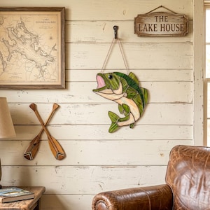 A stained glass fish wall hanging featuring shades of green and cream with a pink mouth, suspended by a metal chain from a black hook. The fish is depicted in a leaping pose with detailed dark solder lines defining its scales and fins.