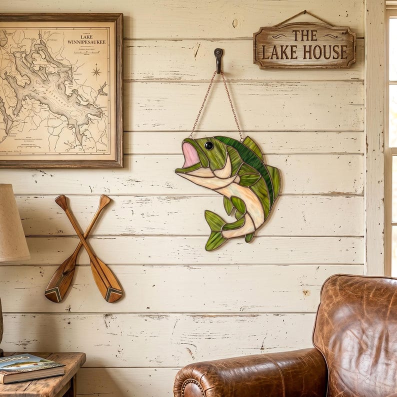 A stained glass fish wall hanging featuring shades of green and cream with a pink mouth, suspended by a metal chain from a black hook. The fish is depicted in a leaping pose with detailed dark solder lines defining its scales and fins.
