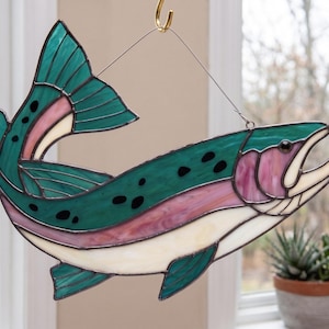 May include: A stained glass suncatcher shaped like a trout fish, featuring deep teal, iridescent pink, and cream-colored glass segments joined by dark metal solder lines, with black spots and a wire hanger for display.