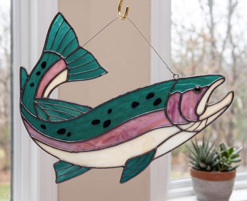 May include: A stained glass suncatcher shaped like a trout fish, featuring deep teal, iridescent pink, and cream-colored glass segments joined by dark metal solder lines, with black spots and a wire hanger for display.