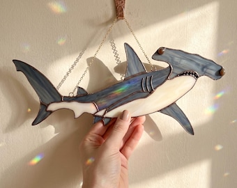 May include: A stained glass hammerhead shark suncatcher featuring translucent blue-grey and cream-colored glass panels joined by dark metal solder lines. The shark is suspended by a thin gold-toned metal chain attached to a copper-colored wall hook.