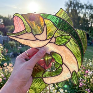 A stained glass suncatcher shaped like a leaping fish, featuring translucent green and cream-colored glass panels held together by dark metal solder lines. The fish has an open mouth and a dark circular eye, with a hanging chain attached.