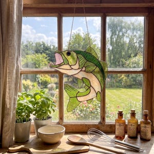 May include: A stained glass suncatcher shaped like a leaping fish with an open mouth. The fish features translucent green and cream-colored glass panels held together by dark metal solder lines. It hangs from a thin metal chain in front of a window.