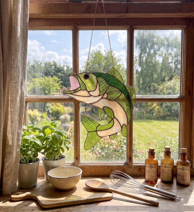 May include: A stained glass suncatcher shaped like a leaping fish with an open mouth. The fish features translucent green and cream-colored glass panels held together by dark metal solder lines. It hangs from a thin metal chain in front of a window.