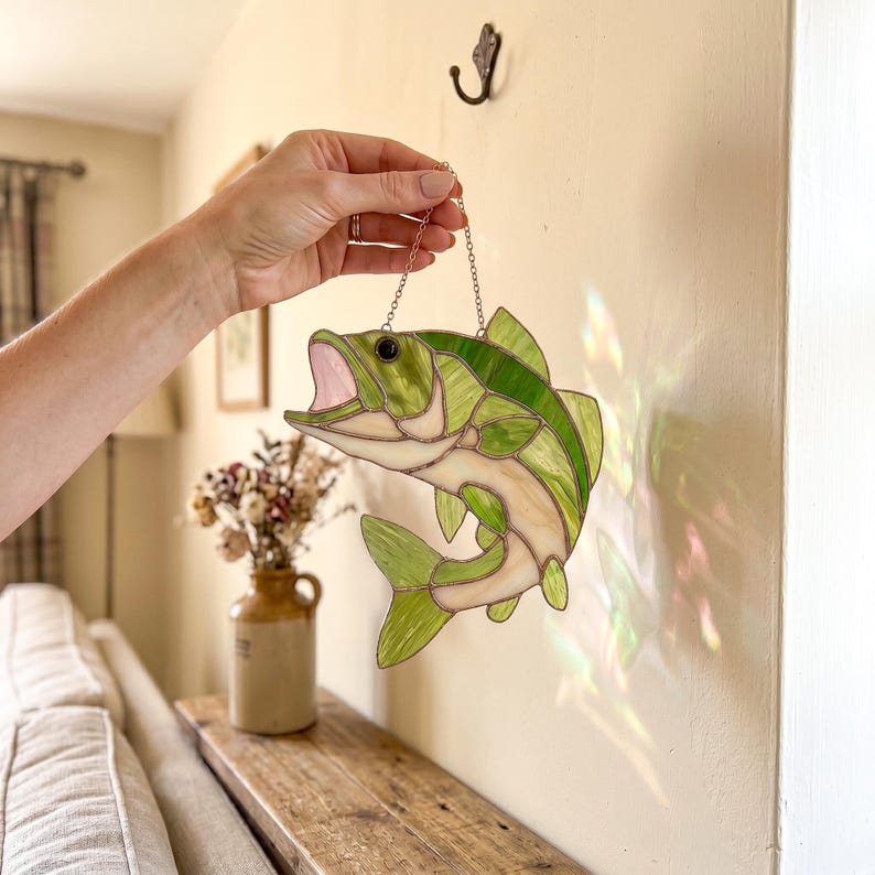 A stained glass suncatcher shaped like a leaping bass fish, featuring various shades of green and cream glass pieces joined by dark metal solder lines. The fish has an open mouth, a black eye, and hangs from a thin metal chain.