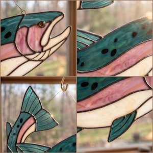 May include: A stained glass suncatcher shaped like a trout, featuring teal, pink, and cream-colored glass pieces joined by dark metal solder lines. The fish design includes a dark eye, black spots, and a hanging wire for display purposes.