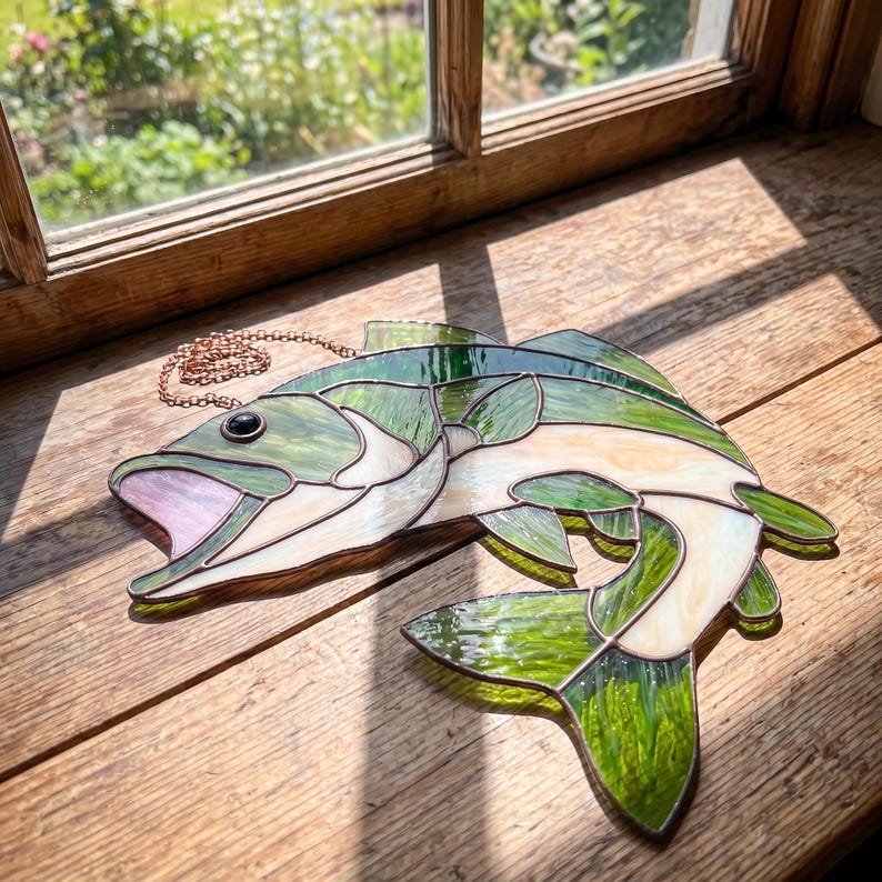 A stained glass fish suncatcher featuring green and cream-colored glass panels joined by copper foil solder lines. The fish has an open mouth with a pink glass interior, a single black glass eye, and an attached copper chain for hanging.