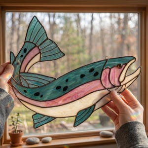 A stained glass suncatcher shaped like a trout, featuring iridescent teal, pink, and cream-colored glass panels joined by dark metal solder lines. The fish design includes black spots along the back and a curved, open-mouthed pose.