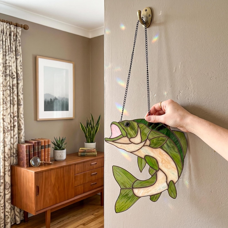 May include: A stained glass suncatcher shaped like a leaping bass fish, featuring variegated green and cream-colored glass panels joined by dark metal solder lines, suspended from a thin black metal chain attached to a wall-mounted hook.